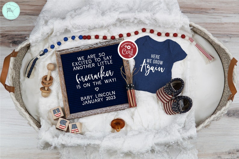 May include: A blue and white letter board with the text "We are so excited to say another little firecracker is on the way! Baby Lincoln January 2023". A blue baby onesie with the text "Here we grow again" is next to the letter board. A red, white, and blue baby rattle, a red, white, and blue beaded necklace, and a red, white, and blue baby shoe are also in the image.