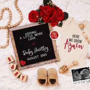 K&ouml;nnte beinhalten: Eine Buchstabentafel mit dem Text "Adding a little more love" und "Baby Hartley August 2023". Ein Baby-Strampler mit dem Text "Here we grow again". Eine Holzrassel, ein Paar Baby-Mokassins und ein gerahmtes Ultraschallbild mit dem Text "Love at first sight".