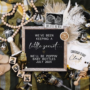 May include: A black letter board with the text "We've been keeping a little secret... We'll be poppin' baby bottles July 202X". A white onesie with the text "Countdown to baby Clark" and a gold glitter firework design. A wooden bead garland, a gold glitter party hat, a baby bottle with a gold glitter bow, and a wooden rattle.