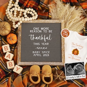 K&ouml;nnte beinhalten: Eine Baby-Ank&uuml;ndigung im Herbst-Stil mit einer Buchstaben-Tafel, auf der steht "One more reason to be thankful this year Baby Spice April 2023". Es gibt Holzbl&ouml;cke, die "BABY" buchstabieren, ein Paar braune Baby-Mokassins, einen kleinen wei&szlig;en Strampler mit einem Truthahn-Motiv und dem Text "The Turkey isn't the only thing in the oven" und ein gerahmtes Ultraschallbild.