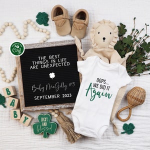 May include: A white onesie with green text that reads "Oops... We Did It Again". The onesie is surrounded by a variety of green shamrocks, a wooden rattle, a stuffed lion, and a letter block spelling "Baby". A black letter board with white text reads "The Best Things In Life Are Unexpected Baby McGilly #3 September 2023".