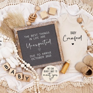May include: A letter board with the text "The best things in life are Unexpected" and "Due to arrive October 2023". A white crocheted teddy bear, wooden blocks spelling "Baby", a wooden sign with the text "We can bearly wait to meet you", a beige onesie with the text "Baby Crawford", and wooden toys are arranged on a white blanket with fringe.