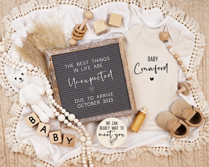 May include: A letter board with the text "The best things in life are Unexpected" and "Due to arrive October 2023". A white crocheted teddy bear, wooden blocks spelling "Baby", a wooden sign with the text "We can bearly wait to meet you", a beige onesie with the text "Baby Crawford", and wooden toys are arranged on a white blanket with fringe.