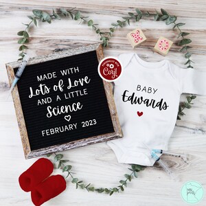 May include: A black letter board with the text "MADE WITH Lots of Love AND A LITTLE Science FEBRUARY 2023". A white onesie with the text "BABY Edwards" and a red heart. Two red booties and two wooden blocks with the letters "X" and "O" are also in the image.