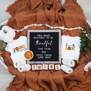 May include: Two white baby grows with Thanksgiving-themed designs, one saying "Double the Blessings" and the other saying "Twice the Love". A letter board with the text "Two more reasons to be thankful this year. Spice Twins June 20XX".  The baby grows and letter board are in a wicker basket with a brown blanket and a garland of wooden beads.