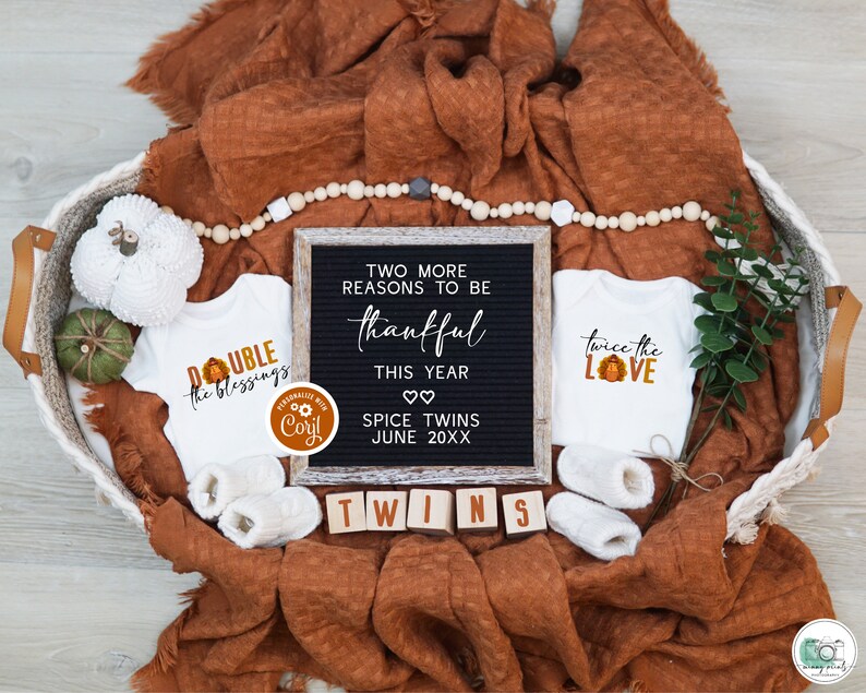 May include: Two white baby grows with Thanksgiving-themed designs, one saying "Double the Blessings" and the other saying "Twice the Love". A letter board with the text "Two more reasons to be thankful this year. Spice Twins June 20XX".  The baby grows and letter board are in a wicker basket with a brown blanket and a garland of wooden beads.