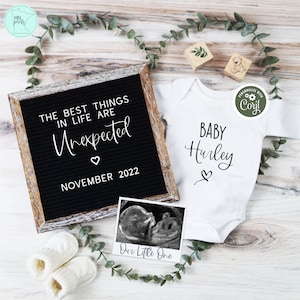 May include: A white onesie with the text "BABY Hurley" and a small heart. A black letter board with the text "THE BEST THINGS IN LIFE ARE Unexpected NOVEMBER 2022". A black and white ultrasound image in a white frame with the text "Our Little One".