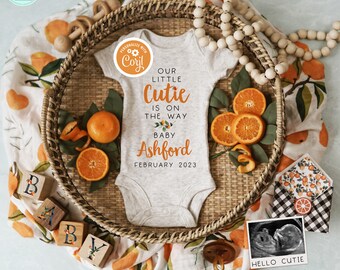 May include: A white babygrow with the text "Our little cutie is on the way baby Ashford February 2023". The babygrow is surrounded by orange slices, wooden blocks that spell "baby", a wooden rattle, and a black and white ultrasound image with the text "Hello cutie".