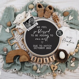 May include: A black letter board with the text "We feel so blessed to be adding to our nest. Due to hatch October 2023."  A white onesie with the text "Baby Hendrix" is next to the letter board.  The onesie is surrounded by wooden beads, a wooden rainbow, and a small wooden cross.