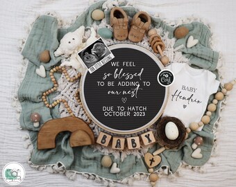 May include: A black letter board with the text "We feel so blessed to be adding to our nest. Due to hatch October 2023."  A white onesie with the text "Baby Hendrix" is next to the letter board.  The onesie is surrounded by wooden beads, a wooden rainbow, and a small wooden cross.