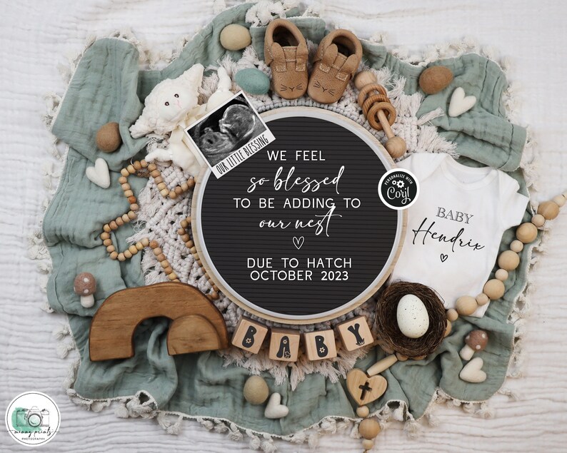 May include: A black letter board with the text "We feel so blessed to be adding to our nest. Due to hatch October 2023."  A white onesie with the text "Baby Hendrix" is next to the letter board.  The onesie is surrounded by wooden beads, a wooden rainbow, and a small wooden cross.
