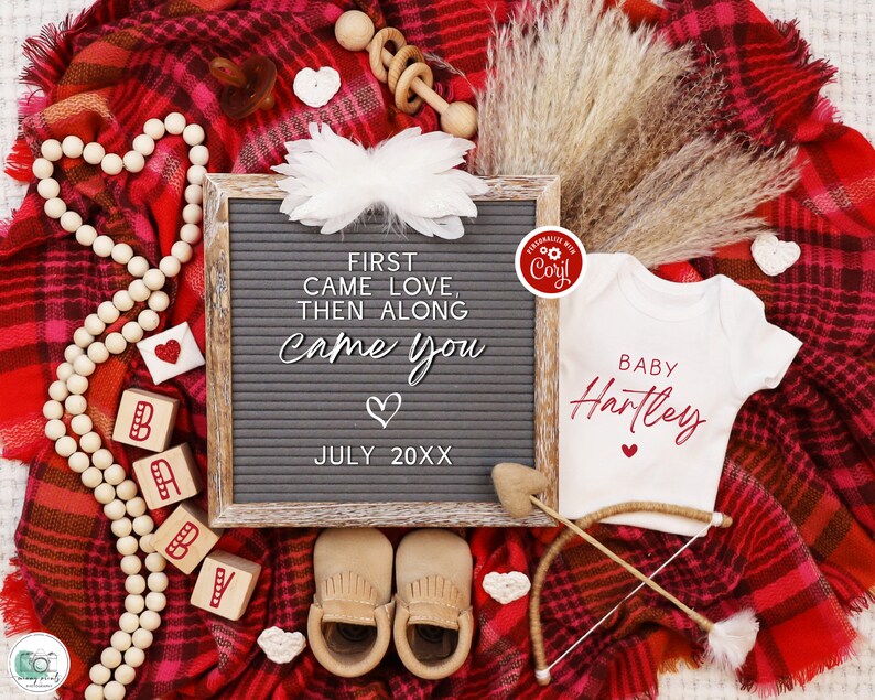 K&ouml;nnte beinhalten: Eine rot-wei&szlig; karierte Decke mit einem wei&szlig;en Strampler, auf dem "BABY Hartley" in roter Schrift steht. Es gibt eine Buchstabentafel, auf der "FIRST CAME LOVE, THEN ALONG CAME YOU" mit einem Herz und "JULY 20XX" steht. Es gibt Holzbl&ouml;cke, die "BABY" buchstabieren, und eine Holzrassel.