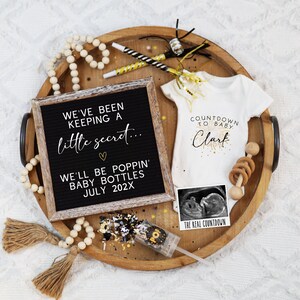 May include: A wooden tray holds a baby announcement. A black letter board reads "We've been keeping a little secret... We'll be poppin' baby bottles July 202X." A white onesie says "Countdown to Baby Clark." Includes a sonogram and confetti.