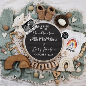 May include: A black chalkboard sign with the text "Blessed with our rainbow but will never forget the storm" and "Baby Hendrix October 2023". A white onesie with a rainbow and the text "After every storm comes a rainbow". A wooden rainbow arch, wooden blocks spelling "baby", and a wooden cross.