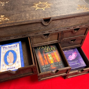 May include: A dark brown wooden box with multiple drawers, each containing oracle cards. The box is decorated with etched floral and sunburst designs. The drawers contain cards labeled "Palmistry," "Past Life," and "Truth."