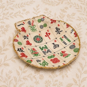 May include: A shallow, shell-shaped dish crafted from a mosaic of white mahjong tiles. The tiles feature various traditional red, green, and black characters and symbols. The scalloped edges of the dish are finished with a metallic gold trim.