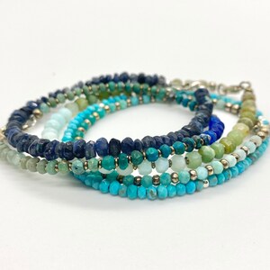 May include: A multi-strand bracelet featuring a gradient of blue, green, and turquoise gemstone beads. The bracelet is made with a variety of different stones, including lapis lazuli, turquoise, and aquamarine.
