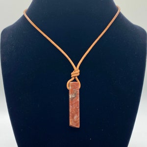 May include: A brown leather cord necklace with a reddish-brown stone pendant. The pendant is a long, thin rectangle with a slightly rounded top and bottom.