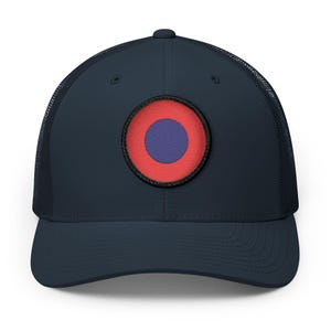 May include: A navy blue trucker hat featuring a mesh back and a circular patch on the front. The patch displays a solid purple center circle surrounded by a wider red ring, all enclosed within a thin black border stitched onto the cap.
