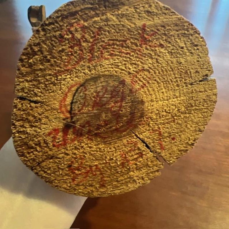 May include: Close-up of a cross-section of a wooden log, showcasing concentric growth rings and a central hole. The wood has a light brown color with visible cracks. Red markings are visible on the surface, possibly handwritten text.