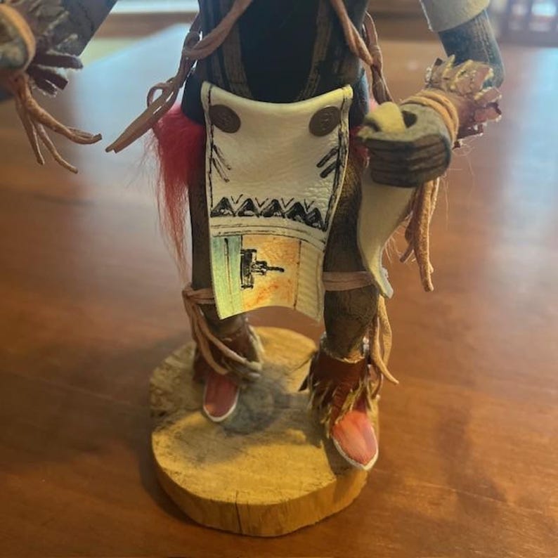 May include: A handcrafted Native American doll with intricate details. The doll features a wooden base, leather clothing, and decorative elements. The doll's attire includes a white apron with geometric designs and a red feather accent.
