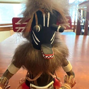 May include: A handcrafted Native American kachina doll with a black face, white markings, and a fur collar. The doll features a red and white headdress and is adorned with feathers. The arms and hands are detailed with painted designs, showcasing traditional craftsmanship.