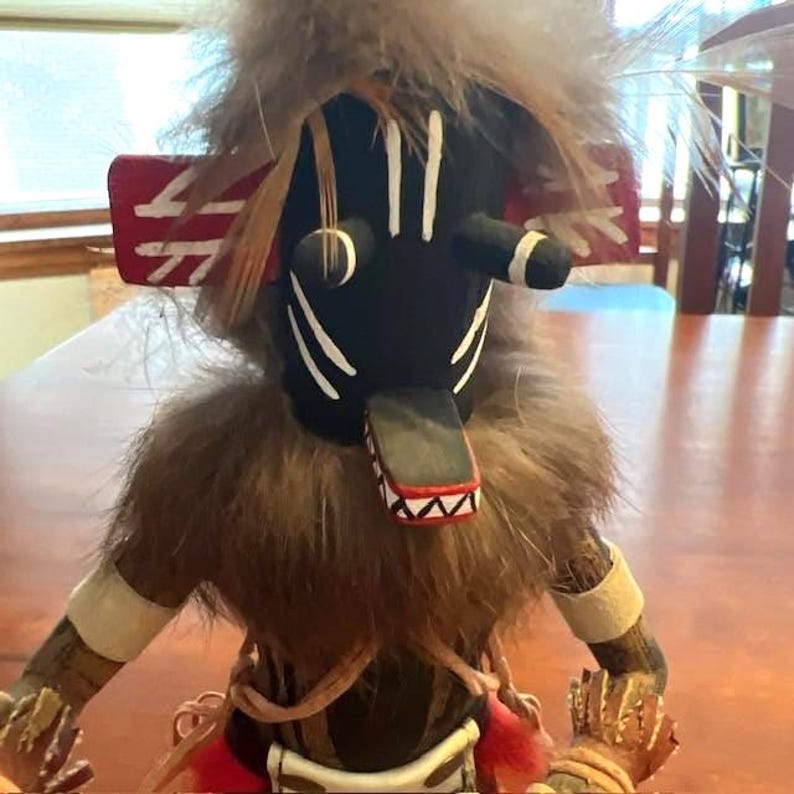 May include: A handcrafted Native American kachina doll with a black face, white markings, and a fur collar. The doll features a red and white headdress and is adorned with feathers. The arms and hands are detailed with painted designs, showcasing traditional craftsmanship.