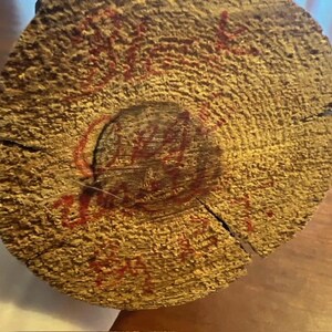 May include: Close-up of a cross-section of a tree trunk, showcasing the wood's texture and growth rings. Red markings are visible on the surface, possibly handwritten text or a stamp. The wood has a light brown color.