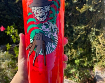 May include: A red, clear plastic tumbler with a green straw. The tumbler has a graphic of Freddy Krueger from the movie "A Nightmare on Elm Street" wearing a red and green striped sweater and holding a glove with blades. The tumbler has a green Starbucks logo and the text "Don't Fall Asleep" on the bottom.