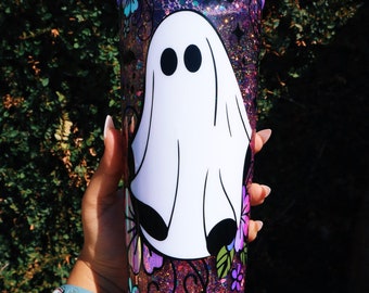 May include: A clear tumbler with a pink, purple, and black glitter design. The tumbler features a white ghost with black eyes and a floral design.