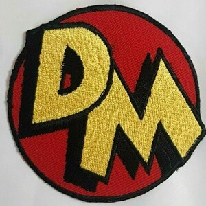 May include: A red, round embroidered patch with the letters "DM" in black and yellow. The letters are stylized and have a cartoonish look.