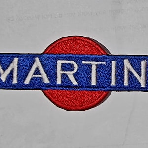 Könnte beinhalten: Ein roter und blauer gestickter Aufnäher mit dem Wort "MARTINI" in weißen Buchstaben.