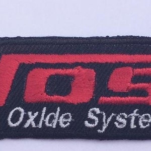 Könnte beinhalten: Ein gestickter schwarzer und roter Aufnäher mit dem Wort "NOS" in Rot und dem Text "Nitrous Oxide Systems" in Weiß darunter. Das "S" in NOS ist als Pfeil nach rechts stilisiert.