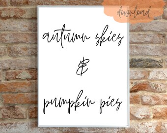 May include: A framed print with the words "autumn skies & pumpkin pies" in black script. The words are centered on a white background. The phrase "instant download" is in a peach-colored bubble in the upper right corner. The background is a brick wall.