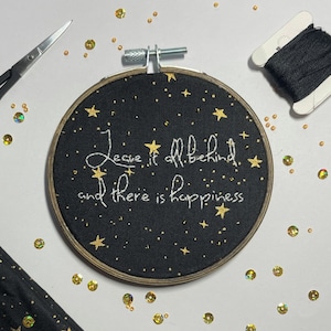 Puede incluir: Un aro de bordado negro con estrellas doradas y el texto "Leave it all behind and there is happiness" bordado en hilo blanco.