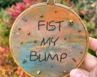 May include: Round embroidery hoop with the text "FIST MY BUMP" stitched in black thread. The fabric has a watercolor effect with orange, blue, and brown hues. Small beads are scattered around the text. The hoop is held by a hand.