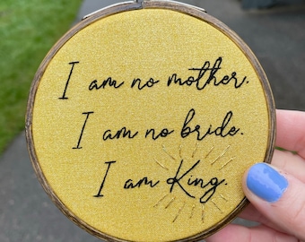 May include: A circular wooden embroidery hoop containing mustard yellow fabric with black embroidered text that reads: I am no mother. I am no bride. I am King. The word King features small gold stitched rays radiating outward from the letters.