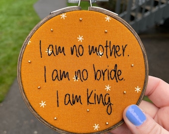 May include: An embroidered hoop art piece with the text "I am no mother. I am no bride. I am King" in black thread on an orange fabric. The design includes small white star and silver bead accents, set in a wooden hoop.