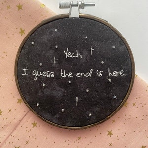 May include: A round, black embroidery hoop with the text "Yeah, I guess the end is here" stitched in white thread. The design includes small crosses and scattered clear beads, set against a starry background. The hoop is framed in wood.