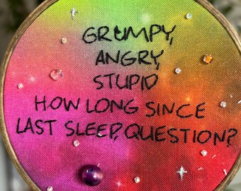 May include: A circular wooden embroidery hoop containing fabric with a vibrant rainbow gradient background. Black embroidered text reads: GRUMPY, ANGRY, STUPID HOW LONG SINCE LAST SLEEP, QUESTION? The fabric is decorated with small clear and purple beads.
