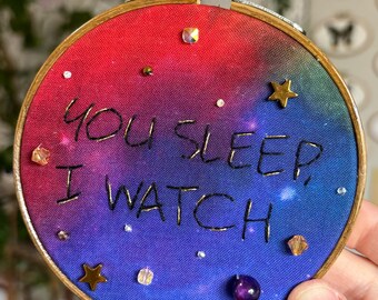 May include: A circular wooden embroidery hoop containing fabric with a red, purple, and blue gradient background. The text "YOU SLEEP, I WATCH" is embroidered in black thread. The piece is decorated with small gold star charms and beads.