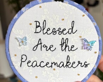May include: A circular embroidery hoop frame wrapped in light blue fabric with a white floral pattern. The center features white fabric with the black embroidered text Blessed Are the Peacemakers, accented by two small translucent butterfly charms.