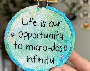 May include: A circular embroidery hoop containing fabric with a teal and green watercolor-style background. The text "Life is our opportunity to micro-dose infinity" is stitched in black thread, accented by small, scattered silver sequins and beads.