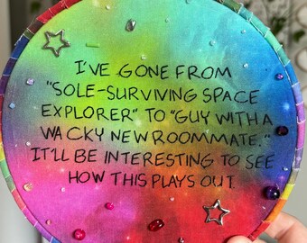 May include: A circular embroidery hoop containing a vibrant, multi-colored tie-dye fabric with black embroidered text that reads: I'VE GONE FROM "SOLE-SURVIVING SPACE EXPLORER" TO "GUY WITH A WACKY NEW ROOMMATE." IT'LL BE INTERESTING TO SEE HOW THIS PLAYS OUT.