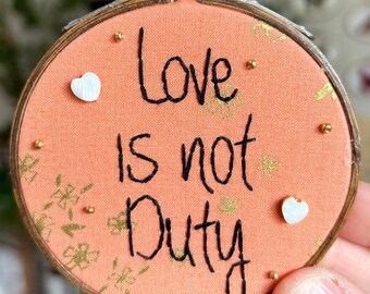May include: A round wooden embroidery hoop containing peach-colored fabric with a gold floral pattern. The black embroidered text reads Love is not Duty. The piece is decorated with two small white heart-shaped buttons and several tiny gold beads.