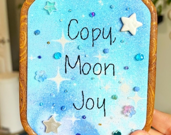 May include: An octagonal wooden embroidery hoop frame containing a light blue fabric background with a watercolor effect. The text "Copy, Moon, Joy" is embroidered in black thread, accented with scattered white star shapes and small blue beads.