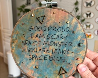 May include: A circular embroidery hoop containing fabric with a mottled teal and copper finish. The fabric features black embroidered text that reads: GOOD PROUD. I AM SCARY SPACE MONSTER. YOU ARE LEAKY SPACE BLOB. Small metal triangles and beads are attached.
