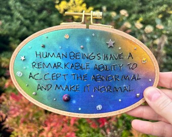 May include: An oval embroidery hoop with a colorful galaxy design. The text reads "HUMAN BEINGS HAVE A REMARKABLE ABILITY TO ACCEPT THE ABNORMAL AND MAKE IT NORMAL." The hoop is decorated with small gems and stars.