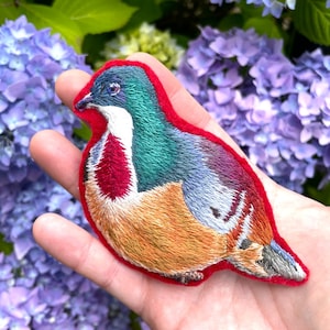Op de afbeelding: Een handgestikte geborduurde vogel broche met een rood, groen, blauw en oranje kleurenschema. De vogel zit op een hand.