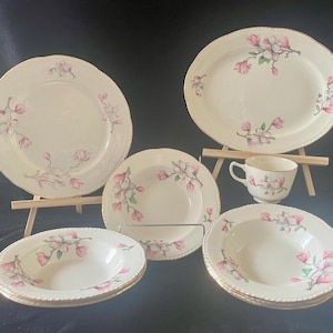 Puede incluir: Un juego de cinco platos de cerámica vintage con estampado floral. Los platos presentan un delicado diseño floral rosa y blanco con ribete dorado. El juego incluye una fuente ovalada grande, dos platos de cena más pequeños, un tazón de sopa y un tazón más pequeño.